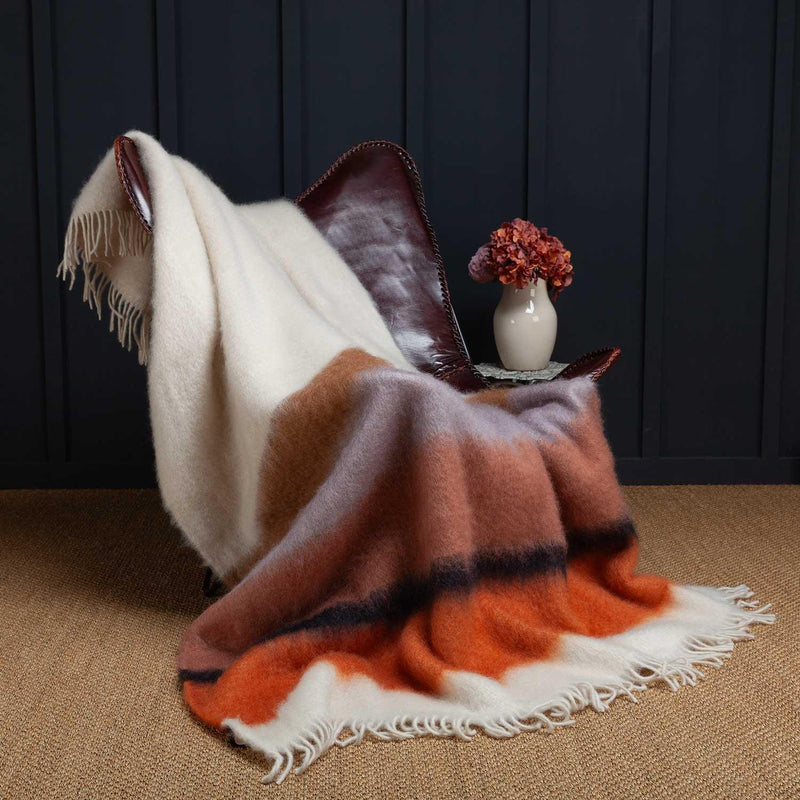 A beautiful blanket woven from a blend of 50% Mohair, 48% Wool, 2% Polyamide with rows of bursts of orange, browns and cream colours, finished with a cream tasselled fringe. Draped over a leather chair. Size 130 x 200 CMS.