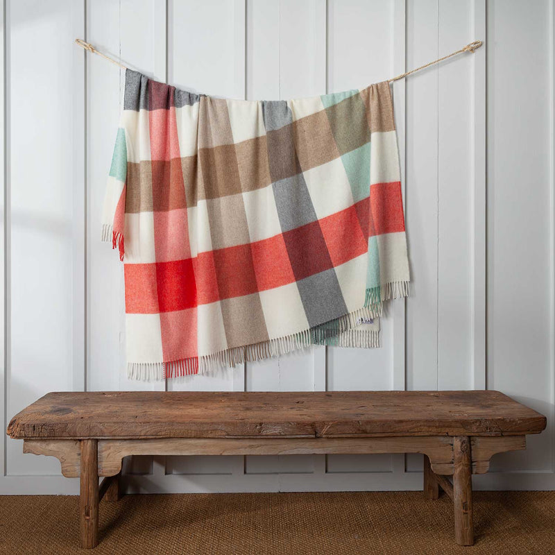 Merino wool block check throw in cream, duck egg, grey, beige and a vibrant coral red. Displayed hanging over a cord on a white wall. 140 x 185 CMS including tassels.