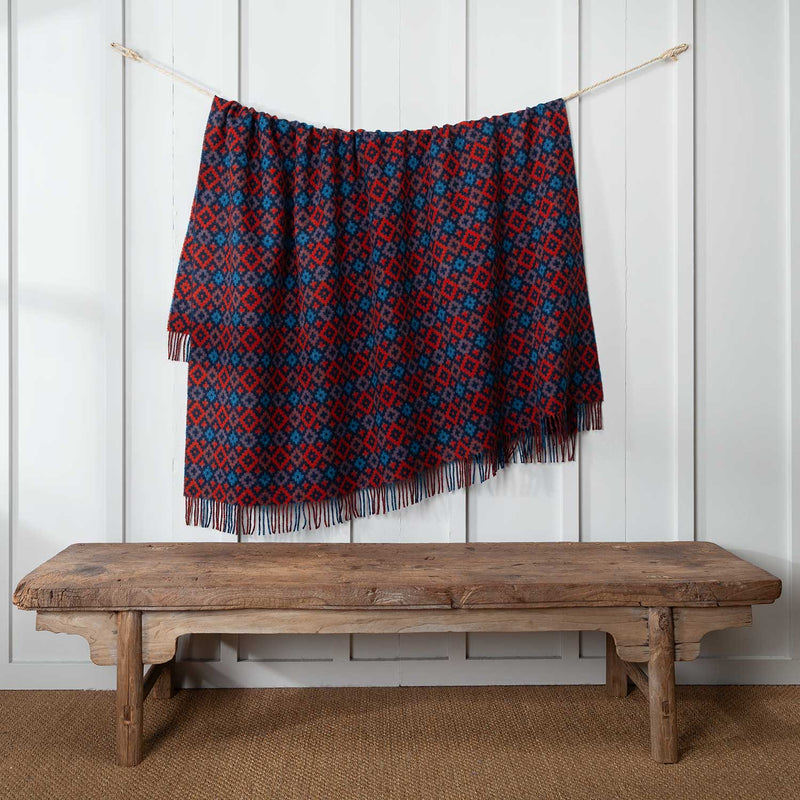 This vibrant wool throw is a rich tapestry of navy, teal and burnt orange in an eye-catching geometric diamond pattern. Displayed on a cord against a white wall.  104 x 185 CMS including tassels.