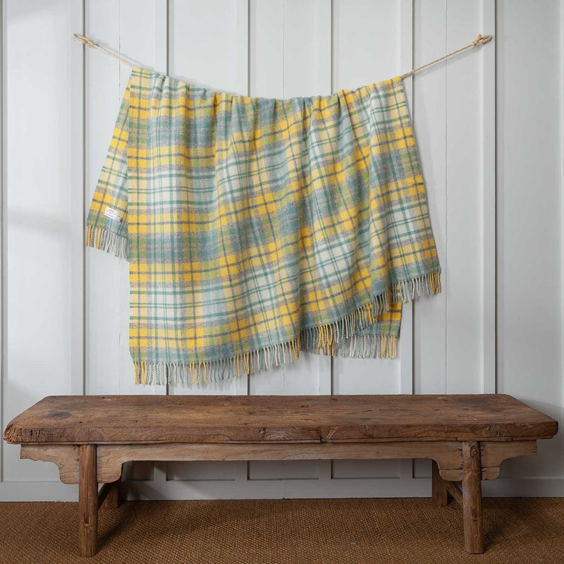 A tartan wool throw in sun-kissed blends of light yellow, soft grey and white. Displayed draped over a cord against a white background.  150 x 183 cm (including tassels).