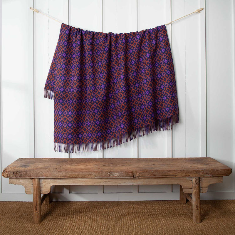 This vibrant wool throw is a rich tapestry of orange, purple, navy and brown in an eye-catching geometric diamond pattern.  Displayed on a cord against a white wall.  104 x 185 CMS including tassels.