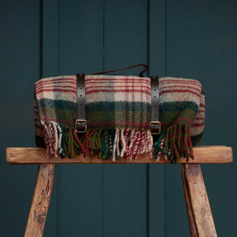 A traditional berry tartan picnic rug in burgundy, forest green, and olive. Complete with a burgundy waterproof backing, brown leather straps and a carry handle. Photographed on a wooden stool. Made in the UK. 100% Pure New Wool.