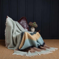 A beautiful blanket woven from a blend of 50% Mohair, 48% Wool, 2% Polyamide. With rows of cool coastal tones of pale green, blue, yellow, and brown, finished with a cream tasselled fringe. Draped over a leather chair. Size 130 x 200 CMS.