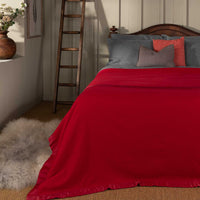 A cherry red Duchess Merino Wool Blanket by John Atkinson with satin binding on a bed withgrey pillows in a room with a wooden ladder and decorative items.  Medium-weight blanket with traditional satin-style ribbon trim.