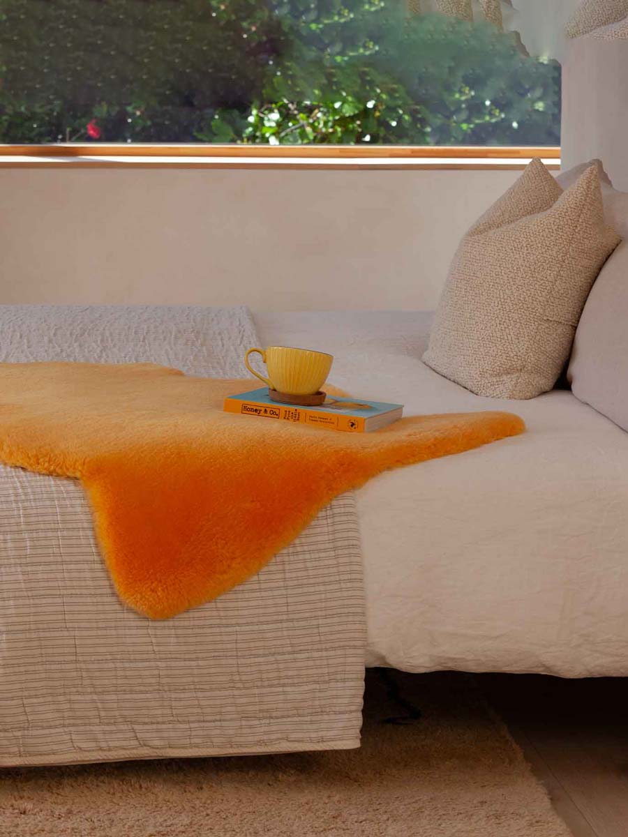 Modern double bed with a bright orange sheepskin rug throw lying on it and a large yellow  cup sitting on a blue and oranges book, with a window behind and greenery outside.