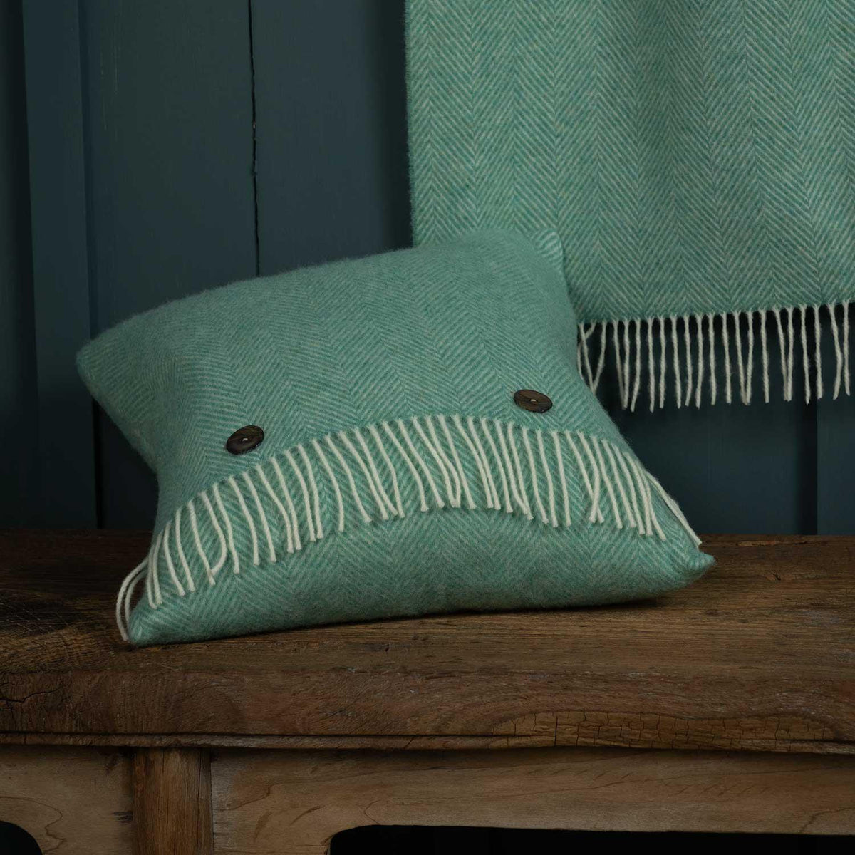 A Merino wool cushion, woven in a herringbone pattern in sea glass green. Contrasting creamy white tassels with 2 mother of pearl buttons. Size 40 x 40 CMS.