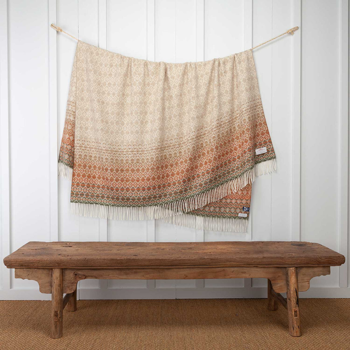 Lambswool throw hung over a cord, beige brown tones with a hint of green at the ends, a diamond pattern, long cream tassels, size is 185 CMS X 140 CMS.