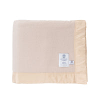 British-made Merino wool blanket in a champagne colour: "Duchess Merino Blanket – Champagne", folded, on a white background, showing the very soft, very light champagne-beige colour of the blanket, the traditional broad satin-style ribbon trim with tidy stitching, the white John Atkinson Duchess" label and the "Woolmark" label.  From The Wool Company, England.