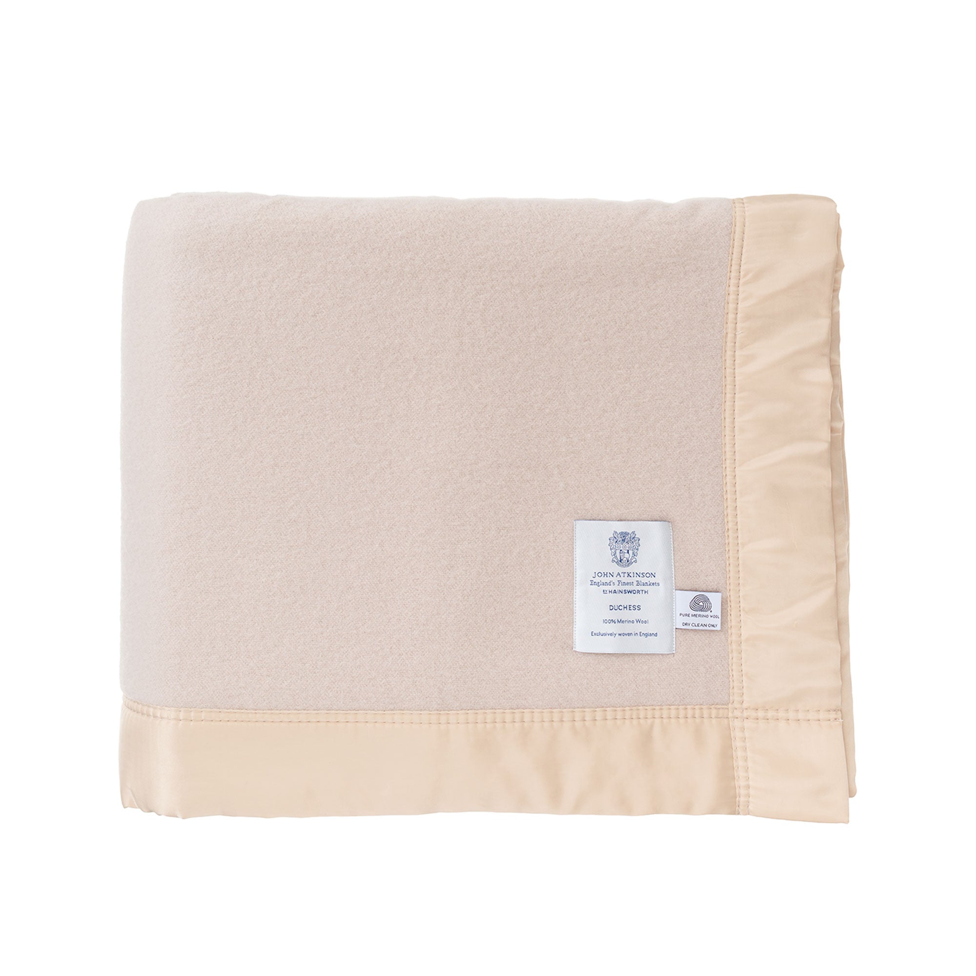 British-made Merino wool blanket in a champagne colour: "Duchess Merino Blanket – Champagne", folded, on a white background, showing the very soft, very light champagne-beige colour of the blanket, the traditional broad satin-style ribbon trim with tidy stitching, the white John Atkinson Duchess" label and the "Woolmark" label.  From The Wool Company, England.