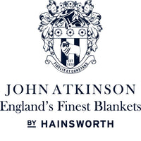 Close-up detail of the blanket label and logo "John Atkinson England's Finest Blankets by Hainsworth", with a coat of arms displaying a white ram over a shield with "Est. 1828", and the motto "Fidelis et Constans", all in blue-black font on a white background.