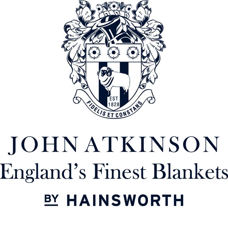 Close-up detail of the blanket label and logo "John Atkinson England's Finest Blankets by Hainsworth", with a coat of arms displaying a white ram over a shield with "Est. 1828", and the motto "Fidelis et Constans", all in blue-black font on a white background.