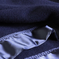 Close-up detail of dark blue "Duchess Merino Blanket - Navy", rucked up to display the deep navy blue of the blanket and the luxuriant sheen of the matching broad satin-style ribbon trim with neat stitching and the detail of the wool fibres. From The Wool Company.